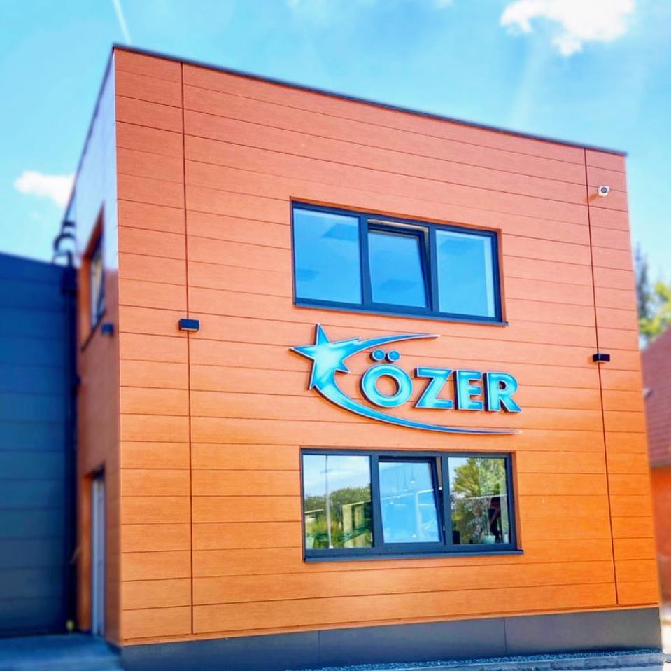 Die Frontseite des Anbaus der Firmenzentrale der Gebäudereinigung Özer GmbH. Ein Holzfarbener rechteckiger moderner Anbau mit vielen Fenstern und dem Özer Logo verziert.