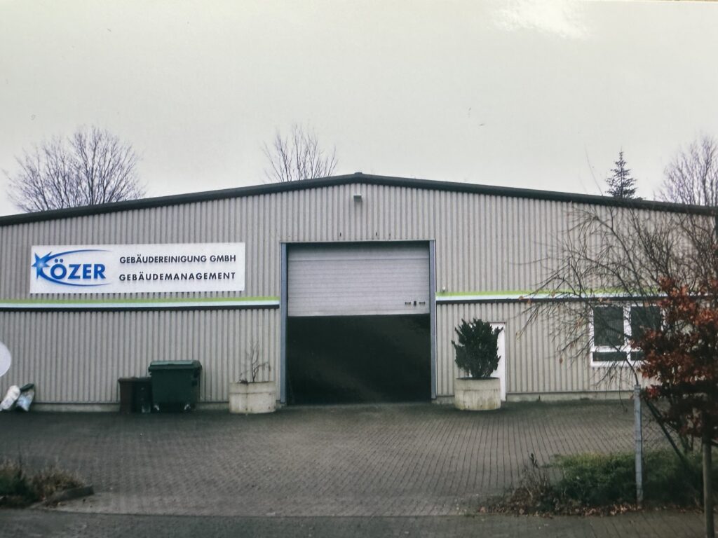 Das Bild zeigt die Zentrale der Gebäudereinigung Özer im Jahr 2000. Eine große graue Lager-Halle mit dem Özer Logo, ein Rolltor ist halb geöffnet.