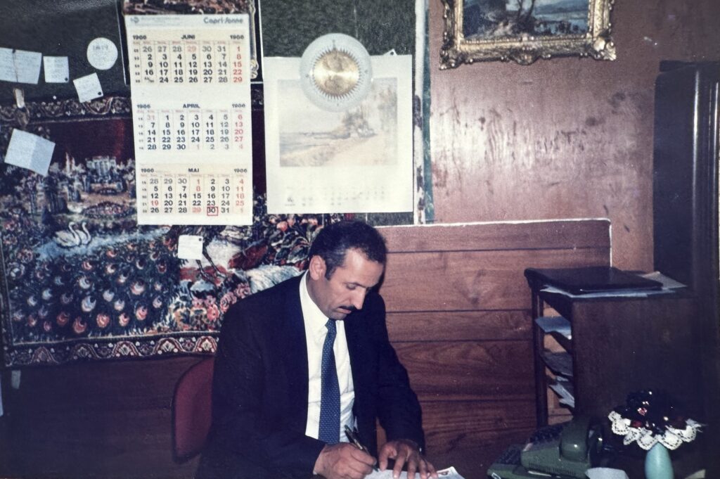 Dunkelhaariger Mann mit Schnauzbart sitzt in seiner Wohnung vor einer Wand mit Holzvertäfelung. Zu sehen ist Firmengründer Ilyas Özer, er trägt einen Anzug, schaut nach unten und schreibt mit einem Stift auf Papier.