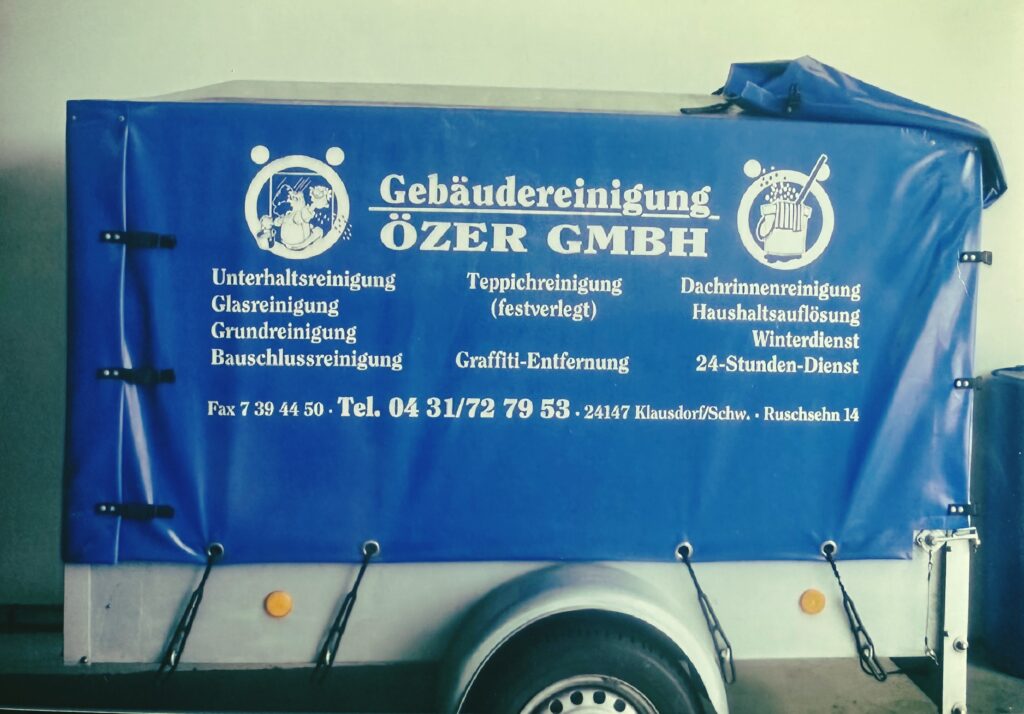 Kleiner Anhänger für Autos mit metallenem Fahrgestellt. Er ist mit einer blauen Plane bespannt, auf der das Özer-Logo, die Leistungen und Telefonnummer zu sehen ist.