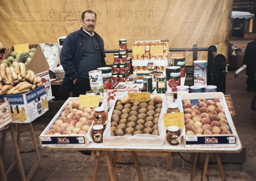Kieler Wochenmarkt 1988. Firmengründer Ilyas Özer steht an seinem Markt-Stand für Obst und Gemüse, zusammen mit türkischen Spezialitäten, die er verkauft. Er trägt einen dunklen Pullover, aus dem ein weißer Hemdkragen herausguckt und eine Jacke. Der Mann hat dunkle Haare, einen Schnauzbart und schaut ernst, aber freundlich in die Kamera.