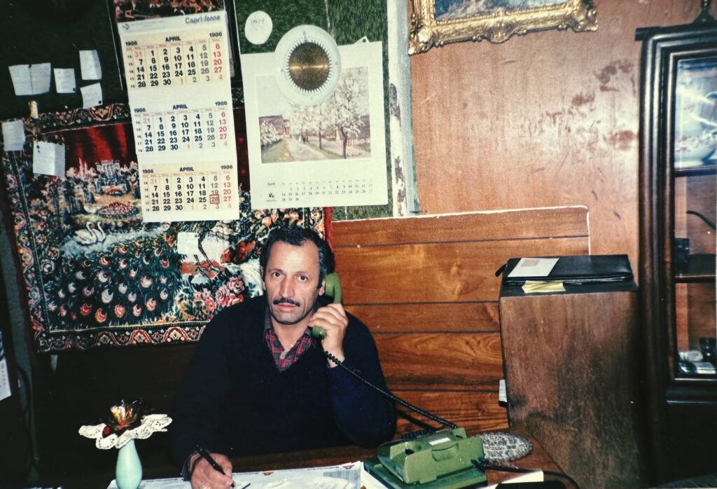 Dunkelhaariger Mann mit Schnauzbart schaut in die Kamera. Zu sehen ist Firmengründer Ilyas Özer sitzt in der 1980er Jahren in seiner Wohnung an einem Tisch, telefoniert mit einem alten grünen Telefon, trägt einen dunkelblauen Pullover, aus dem oben ein rot-brauner Hemdkragen herausguckt.