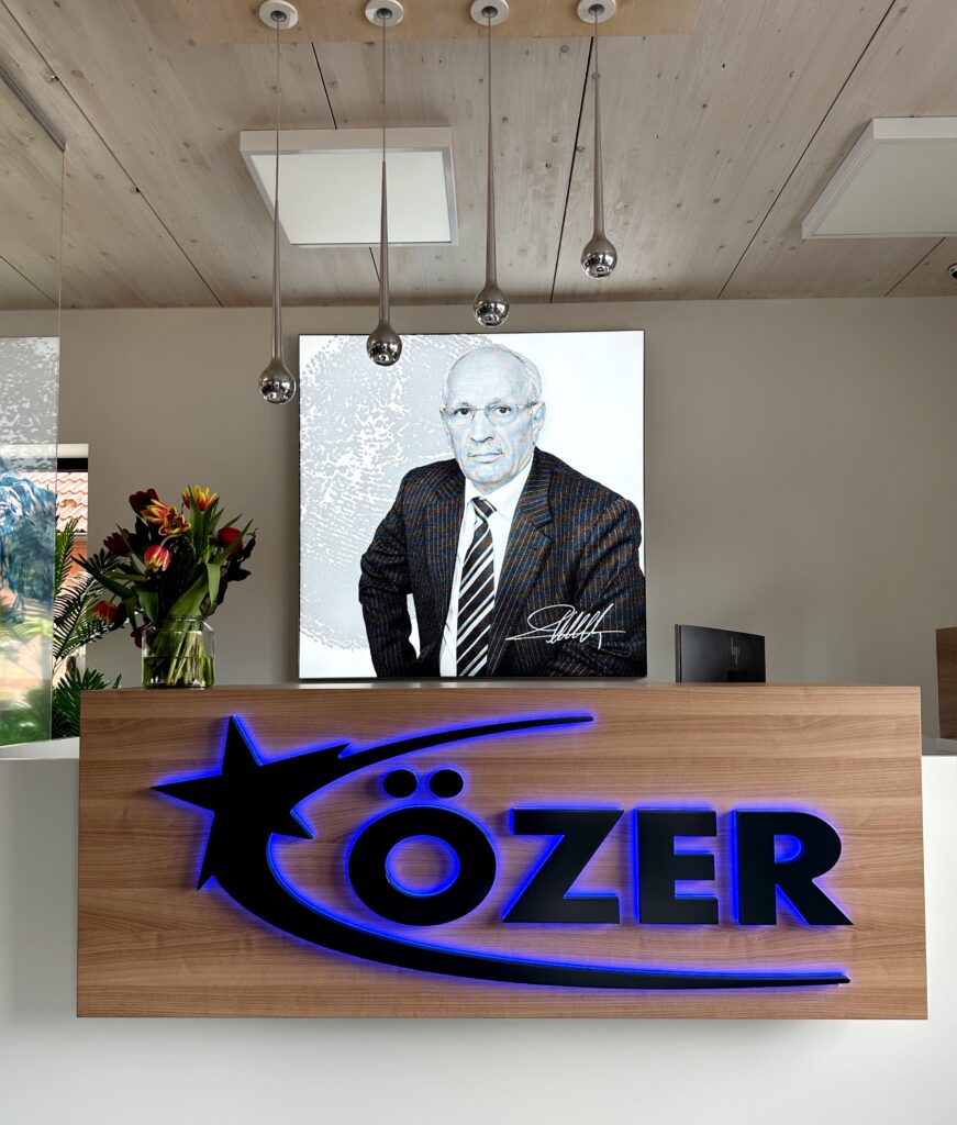 Der Empfangsbereich der Firmenzentrale. Empfangstresen mit dem Özer Logo, Blumen auf dem Tresen, im Hintergrund ein beleuchtetes Porträt des Firmengründers Ilyas Özer.